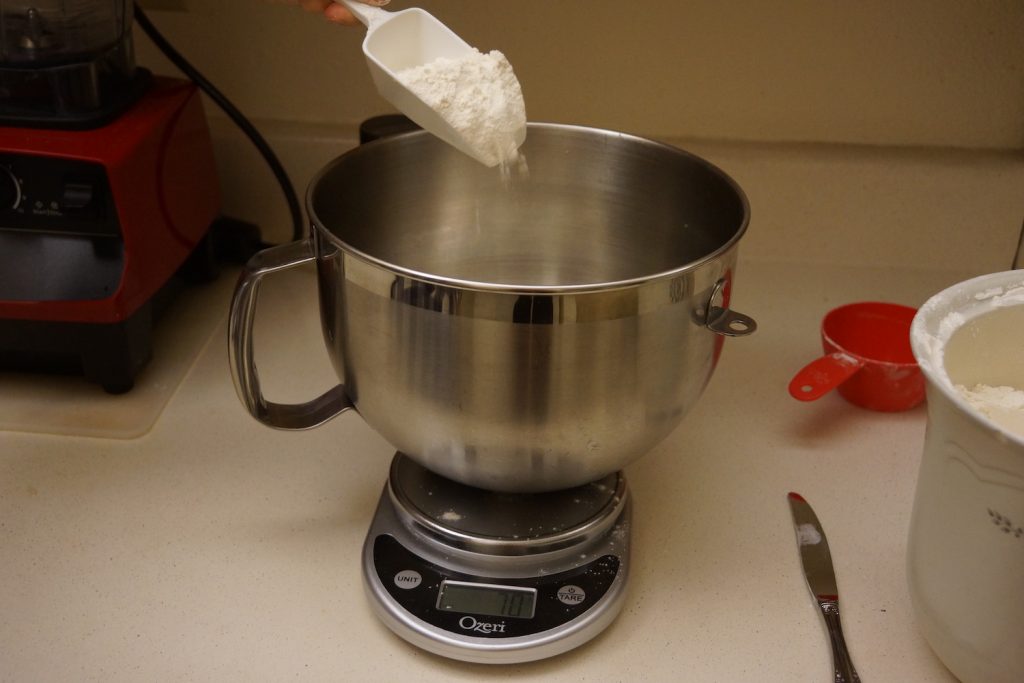 Flour being scooped into a bowl sitting on a kitchen scale